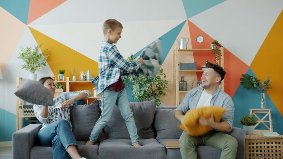 Mother, father and adorable active kid are fighting pillows having fun together on sofa in house enjoying family activity. People, happiness and youth concept.