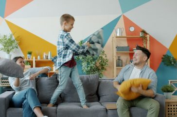 Mother, father and adorable active kid are fighting pillows having fun together on sofa in house enjoying family activity. People, happiness and youth concept.
